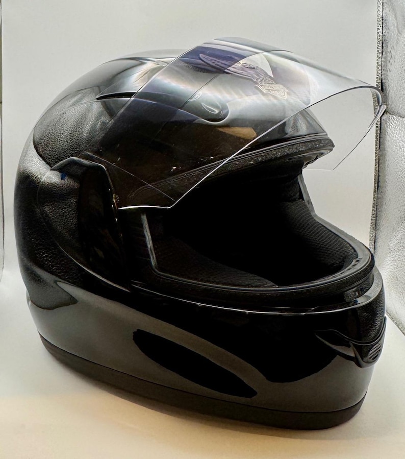 Peut inclure: Casque de moto noir brillant avec une visi&egrave;re transparente relev&eacute;e. Le casque pr&eacute;sente un design profil&eacute; et une surface textur&eacute;e sur le dessus. L'int&eacute;rieur rembourr&eacute; est visible.