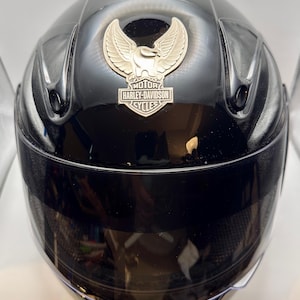 Peut inclure: Casque de moto noir brillant avec visi&egrave;re teint&eacute;e. Le casque pr&eacute;sente un embl&egrave;me argent&eacute; avec les mots "MOTOR HARLEY-DAVIDSON CYCLES" et un motif d'aigle stylis&eacute;.