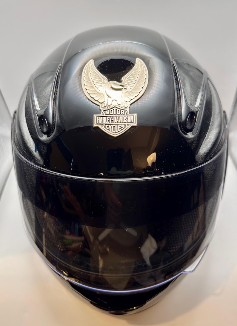 Peut inclure: Casque de moto noir brillant avec visi&egrave;re teint&eacute;e. Le casque pr&eacute;sente un embl&egrave;me argent&eacute; avec les mots "MOTOR HARLEY-DAVIDSON CYCLES" et un motif d'aigle stylis&eacute;.