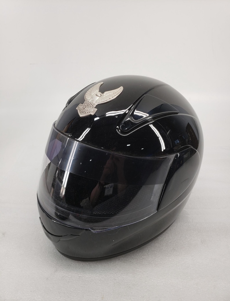 Peut inclure: Casque de moto noir brillant avec visi&egrave;re transparente. Un embl&egrave;me argent&eacute; avec un motif ail&eacute; est fix&eacute; sur le devant. Le casque est con&ccedil;u pour la s&eacute;curit&eacute; et la protection des motocyclistes.