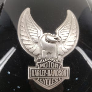 Peut inclure: Gros plan sur un casque de moto noir avec un embl&egrave;me Harley-Davidson argent&eacute;. L'embl&egrave;me repr&eacute;sente un aigle aux ailes d&eacute;ploy&eacute;es au-dessus des mots "MOTOR HARLEY-DAVIDSON CYCLES". Le casque a une finition brillante.