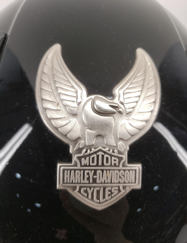 Peut inclure: Gros plan sur un casque de moto noir avec un embl&egrave;me Harley-Davidson argent&eacute;. L'embl&egrave;me repr&eacute;sente un aigle aux ailes d&eacute;ploy&eacute;es au-dessus des mots "MOTOR HARLEY-DAVIDSON CYCLES". Le casque a une finition brillante.