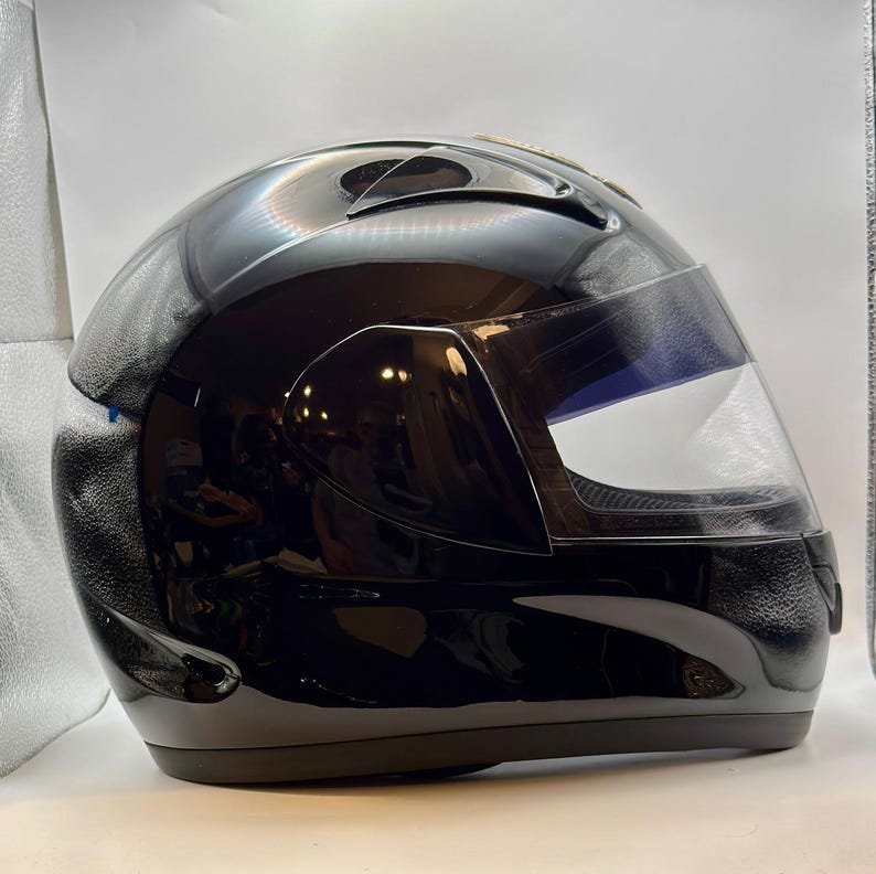 Peut inclure: Un casque de moto noir brillant avec une visi&egrave;re transparente. Le casque a un design &eacute;pur&eacute; avec des ouvertures sur le dessus et un int&eacute;rieur sombre. La visi&egrave;re est l&eacute;g&egrave;rement teint&eacute;e et &eacute;pouse le visage.