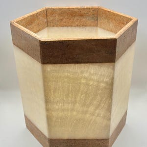 May include: A hexagonal wastebasket featuring alternating horizontal bands of smooth, translucent cream-colored stone and textured, dark brown stone. The vessel has a wide, open top and a solid base, showcasing a clean, geometric design with natural stone variations throughout the exterior surface.