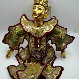 May include: A golden-faced puppet with a conical headdress, adorned with red, green, and gold embellishments. The puppet wears a green and gold costume with red trim and gold sequins. The puppet's hands and feet are also gold.