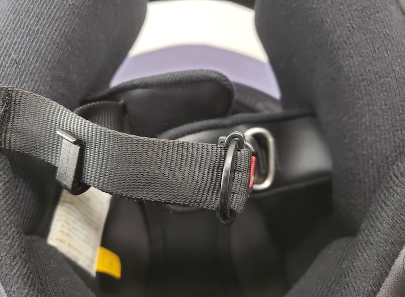 Peut inclure: Gros plan de l'int&eacute;rieur d'un casque de moto noir. L'image met en &eacute;vidence la jugulaire noire avec un anneau en D en m&eacute;tal et un clip en plastique noir. Le rembourrage int&eacute;rieur du casque est visible.