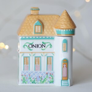 K&ouml;nnte beinhalten: Ein wei&szlig;er, keramik-gefertigter Gew&uuml;rztopf in Form eines Hauses mit einem gelben Dach, gr&uuml;nen Verzierungen und dem Wort "ONION" auf der Vorderseite. Der Topf hat einen kleinen Turm auf der rechten Seite.