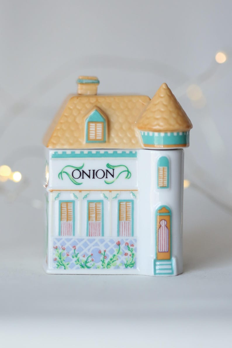 K&ouml;nnte beinhalten: Ein wei&szlig;er, keramik-gefertigter Gew&uuml;rztopf in Form eines Hauses mit einem gelben Dach, gr&uuml;nen Verzierungen und dem Wort "ONION" auf der Vorderseite. Der Topf hat einen kleinen Turm auf der rechten Seite.