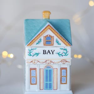 K&ouml;nnte beinhalten: Ein wei&szlig;er, keramik-gefertigter Gew&uuml;rztopf in Form eines Hauses mit einem blauen Dach, gelben Verzierungen und einem gr&uuml;nen Blumendesign. Der Topf ist mit "BAY" beschriftet.