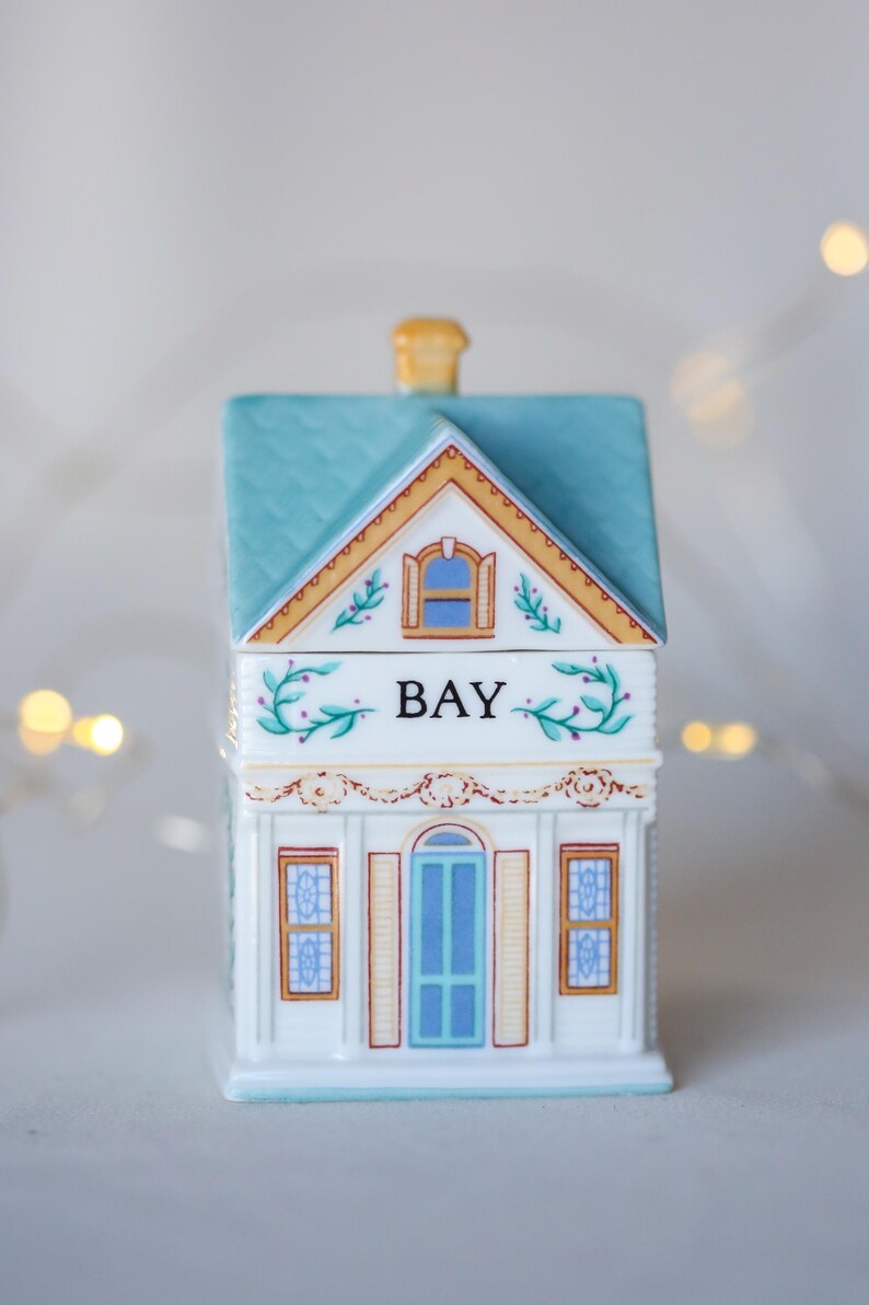 K&ouml;nnte beinhalten: Ein wei&szlig;er, keramik-gefertigter Gew&uuml;rztopf in Form eines Hauses mit einem blauen Dach, gelben Verzierungen und einem gr&uuml;nen Blumendesign. Der Topf ist mit "BAY" beschriftet.