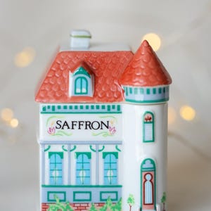 K&ouml;nnte beinhalten: Ein wei&szlig;er, keramik-gefertigter Gew&uuml;rztopf in Form eines Hauses mit rotem Dach, gr&uuml;nem Zierleisten und dem Wort "SAFFRON" auf der Vorderseite. Der Topf hat einen kleinen Schornstein und einen Turm mit einem Fenster.