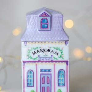 K&ouml;nnte beinhalten: Ein wei&szlig;er Keramik-Gew&uuml;rztopf in Form eines Hauses mit einem violetten Dach und einer rosafarbenen T&uuml;r. Der Topf ist mit einem Blumendesign verziert und das Wort "MARJORAM" steht auf der Vorderseite.
