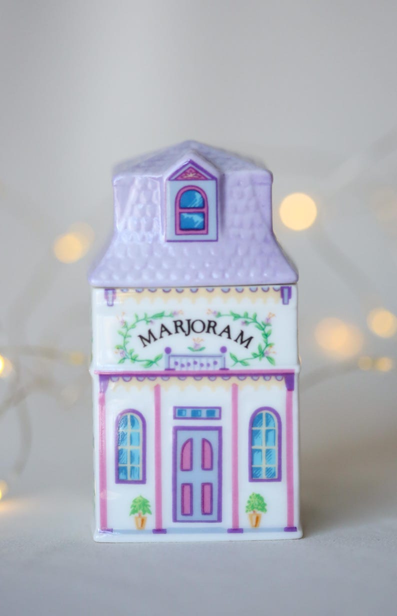 K&ouml;nnte beinhalten: Ein wei&szlig;er Keramik-Gew&uuml;rztopf in Form eines Hauses mit einem violetten Dach und einer rosafarbenen T&uuml;r. Der Topf ist mit einem Blumendesign verziert und das Wort "MARJORAM" steht auf der Vorderseite.