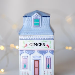 K&ouml;nnte beinhalten: Ein wei&szlig;er Keramik-Gew&uuml;rztopf in Form eines Hauses mit blauem Dach und rosa und blauen Verzierungen. Der Topf hat ein Fenster mit einer gr&uuml;nen Scheibe und steht "GINGER" auf der Vorderseite.