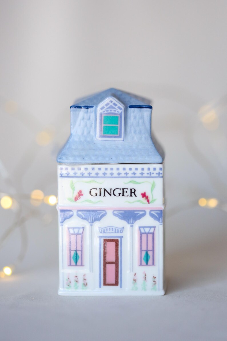 K&ouml;nnte beinhalten: Ein wei&szlig;er Keramik-Gew&uuml;rztopf in Form eines Hauses mit blauem Dach und rosa und blauen Verzierungen. Der Topf hat ein Fenster mit einer gr&uuml;nen Scheibe und steht "GINGER" auf der Vorderseite.