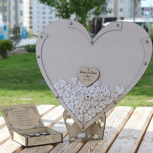 May include: A white wooden heart-shaped guest book with a stand. The heart is filled with small wooden hearts with writing on them. The text on the large heart says "Mary & Charles - May 2023".
