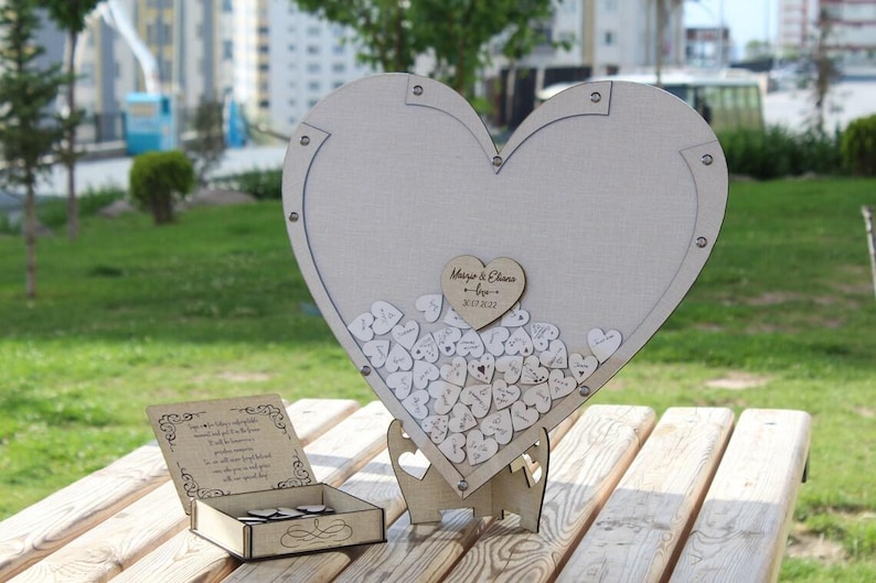 May include: A white wooden heart-shaped guest book with a stand. The heart is filled with small wooden hearts with writing on them. The text on the large heart says "Mary & Charles - May 2023".