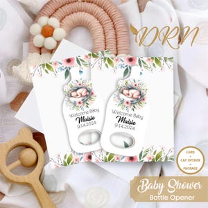 May include: Two white card bottle openers with a floral design and a baby sleeping in a bottle. The text on the card reads "Welcome Baby Maisie 9.14.2024".