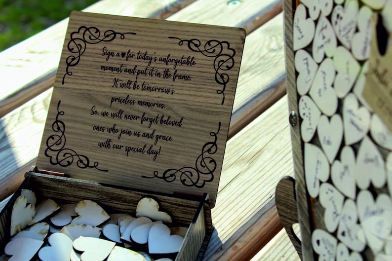 May include: Wooden box with a sign that reads "Sign a heart for today's unforgettable moment and put it in the frame. It will be tomorrow's priceless memories. So, we will never forget beloved ones who join us and grace with our special day!" The box is filled with small wooden hearts.