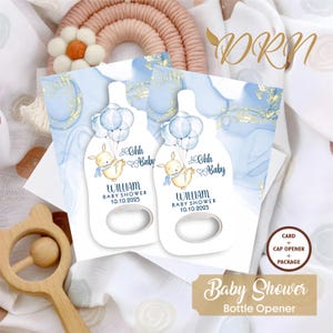 Pode incluir: Dois abridores de garrafas de chá de bebê azul e branco com um coelho fofo segurando balões. Os favores são decorados com detalhes em folha de ouro e o texto "Ohh Baby" e "William Baby Shower 10.10.2025".