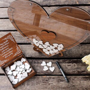 May include: Wooden heart-shaped guest book with a clear acrylic cover. The guest book is filled with white wooden hearts for guests to write messages on. The guest book is sitting on a wooden table with a bouquet of white tulips.