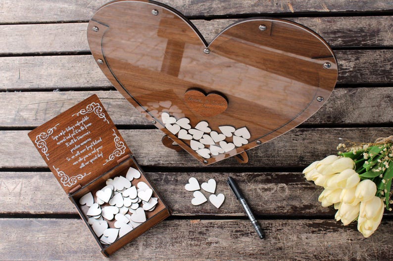 May include: Wooden heart-shaped guest book with a clear acrylic cover. The guest book is filled with white wooden hearts for guests to write messages on. The guest book is sitting on a wooden table with a bouquet of white tulips.