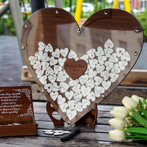 May include: A wooden heart-shaped guest book with a clear acrylic cover. The heart is filled with small wooden hearts that guests can write on. The heart is on a wooden stand. The text on the heart reads "Marie & Paul - Forever".
