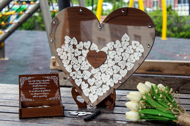 May include: A wooden heart-shaped guest book with a clear acrylic cover. The heart is filled with small wooden hearts that guests can write on. The heart is on a wooden stand. The text on the heart reads "Marie & Paul - Forever".