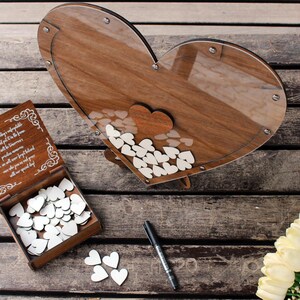 May include: A wooden heart-shaped box with a glass front, filled with small white wooden hearts. The box is on a wooden surface with a small wooden box with a white inscription and a pen next to it. A bouquet of white flowers is in the lower right corner of the image.
