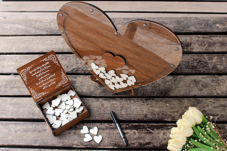 May include: A wooden heart-shaped box with a glass front, filled with small white wooden hearts. The box is on a wooden surface with a small wooden box with a white inscription and a pen next to it. A bouquet of white flowers is in the lower right corner of the image.