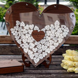 May include: A wooden heart-shaped box with a clear acrylic front, filled with small white wooden hearts for guests to sign at a wedding. The box is on a wooden stand and has a small wooden box with a pen next to it. The text on the box reads "Made for Each Other - Happy Ever After".