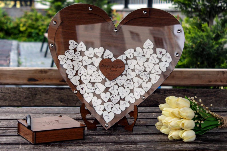 May include: A wooden heart-shaped box with a clear acrylic front, filled with small white wooden hearts for guests to sign at a wedding. The box is on a wooden stand and has a small wooden box with a pen next to it. The text on the box reads "Made for Each Other - Happy Ever After".