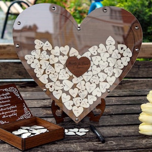 May include: A wooden heart-shaped guest book with a clear acrylic cover. The heart is filled with small wooden hearts that guests can sign. The guest book is on a wooden stand and there is a black pen next to it. The text on the heart reads "Melissa & Steve - August 2023".