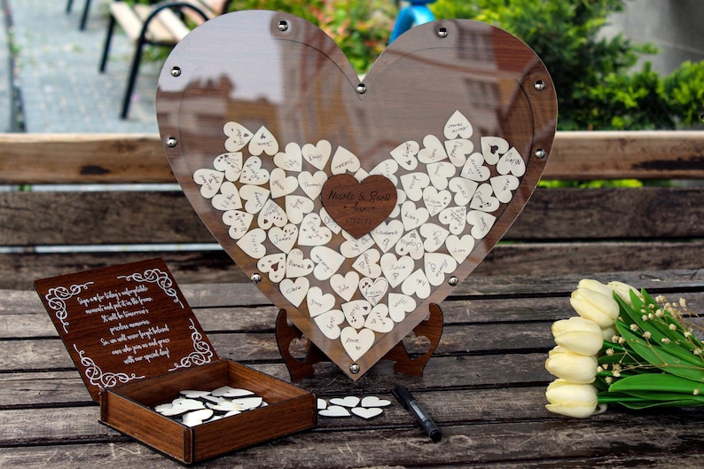 May include: A wooden heart-shaped guest book with a clear acrylic cover. The heart is filled with small wooden hearts that guests can sign. The guest book is on a wooden stand and there is a black pen next to it. The text on the heart reads "Melissa & Steve - August 2023".