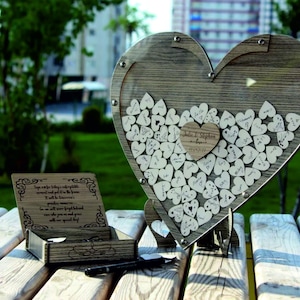 May include: A wooden heart-shaped box with a clear lid filled with small wooden hearts for guests to sign at a wedding. The box is on a wooden table with a small wooden box next to it.