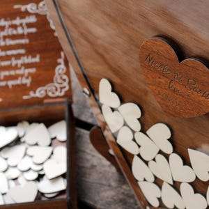 May include: A wooden box with a heart-shaped lid. The lid has the words "Nicole & Scott Love 1.21.23" engraved on it. The box is filled with small white wooden hearts.