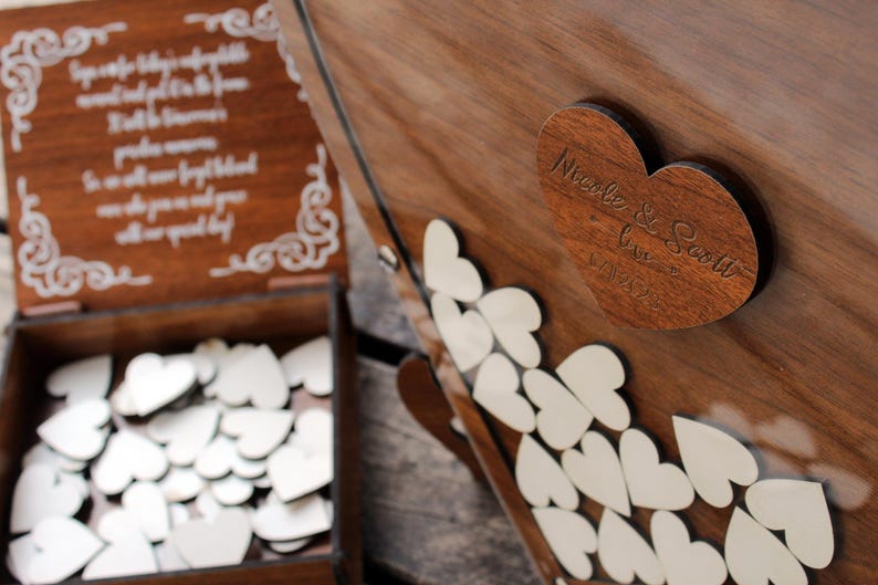 May include: A wooden box with a heart-shaped lid. The lid has the words "Nicole & Scott Love 1.21.23" engraved on it. The box is filled with small white wooden hearts.