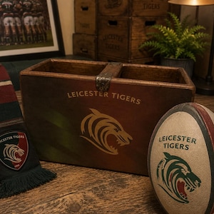 May include: A rectangular wooden storage box featuring the Leicester Tigers logo and text, accompanied by a rugby ball with the same branding and a striped scarf with a team emblem, all displaying the team's signature colours and graphics.