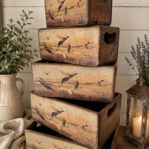 May include: A stack of five rectangular wooden storage crates with cutout handles, each featuring a printed scene of a hunter with dogs and flying pheasants in a field. The boxes have a distressed, aged finish with warm brown and tan tones.