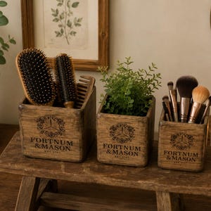 May include: Three wooden boxes, each with the text "FORTNUM & MASON EST. 1707", sit on a small wooden stool. The boxes contain hairbrushes, a plant, and makeup brushes. A lit candle and a mirror are in the background.