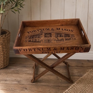 May include: A rectangular wooden serving tray with cutout handles, featuring a dark brown finish and a black graphic illustration of a storefront. The text FORTNUM & MASON EST 1707 is printed on the tray. It rests on a folding wooden X-frame stand.