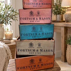 May include: A stack of five rectangular wooden crates in varying colours including pink, blue, cream, and orange. Each crate features the black printed text FORTNUM & MASON EST 1707 along with two crests centred above the brand name on the front panel.