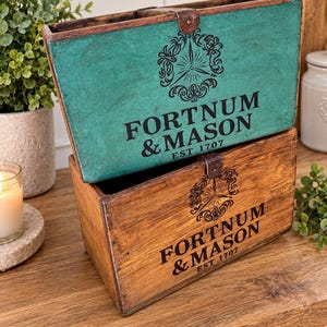 May include: Two rectangular wooden storage crates stacked vertically. The top crate is painted teal with a dark brown metal latch, while the bottom crate is natural wood. Both feature black printed text reading FORTNUM & MASON EST 1707.