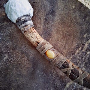 Könnte beinhalten: Ein Trommelschlägel mit einem weiß fellbezogenen Kopf, einem mit grauen Lederstreifen umwickelten Holzgriff und einer zentralen, bernsteinfarbenen ovalen Steineinlage. Der Griff ist mit überkreuzten Lederbändern am unteren Teil befestigt.