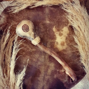 Peut inclure: Un tambour artisanal avec une surface en cuir marron et un batteur en bois. Le batteur présente un motif en spirale et est enveloppé de ficelle. De l'herbe ornementale séchée entoure le tambour, créant une esthétique naturelle et terreuse.