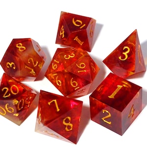 Op de afbeelding: Set van zeven rode en gouden dobbelstenen met de nummers 1-6, 8, 12 en 20. De dobbelstenen zijn gemaakt van helder hars met gouden vlokken en gouden nummers.