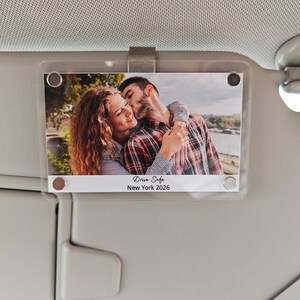Peut inclure: Support photo de pare-soleil de voiture en plastique transparent avec une photo d'un couple s'embrassant. La photo porte les mots "Drive Safe" et "New York 2026". Le support est fixé à un pare-soleil de voiture.