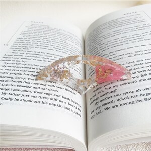 A resin book page holder featuring pink flowers and gold flecks, embedded in clear resin, placed between the pages of an open book. The holder gently holds the pages in place, adding a delicate and elegant touch to the reading experience