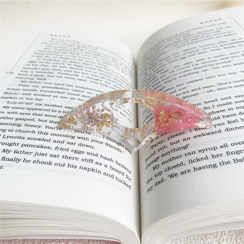 A resin book page holder featuring pink flowers and gold flecks, embedded in clear resin, placed between the pages of an open book. The holder gently holds the pages in place, adding a delicate and elegant touch to the reading experience