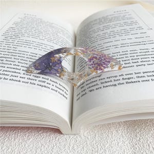 A resin book page holder featuring purple flowers and gold flecks embedded in clear resin, placed between the pages of an open book. The holder gently holds the pages in place, adding an elegant and delicate touch to the reading experience